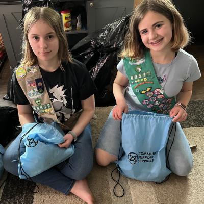 Renee and Amber Dunham wear their Girl Scout sashes and sit on the floor while holding bags that say "Community Support Services."