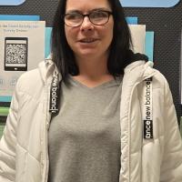 A photo of Amanda, a white woman with black, shoulder-length hair, dark-framed glasses, a white winter coat and a gray T-shirt
