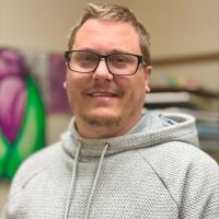 A photo of Zach, a smiling white man with blonde hair and dark-framed glasses wearing a gray hoodie.