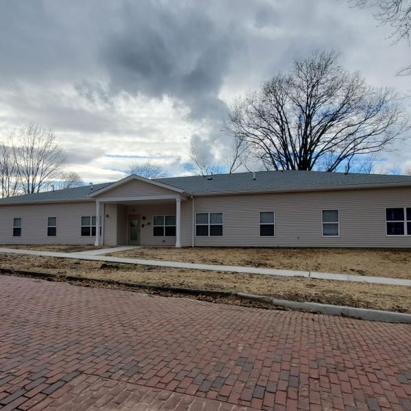 The Naomi Project, a long, one-story tan building, sits below an overcast sky with a brick street stretching in front.