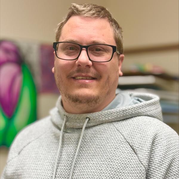 A photo of Zach, a smiling white man with blonde hair and dark-framed glasses wearing a gray hoodie.